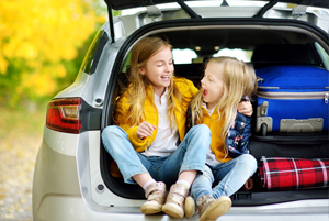 Two young girls sit in the open trunk of a car packed with luggage, laughing and smiling at each other against a backdrop of autumn leaves