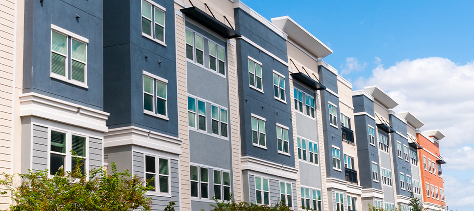 A modern multi-story apartment complex with blue, grey, and tan siding under a clear blue sky