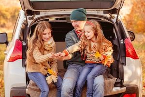 Two young girls sit in the open trunk of a car packed with luggage, laughing and smiling at each other against a backdrop of autumn leaves